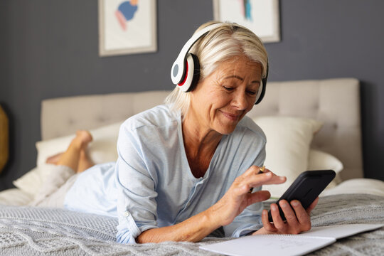 Happy senior caucasian woman laying on bed in bedroom, using smartphone and wearing headphones