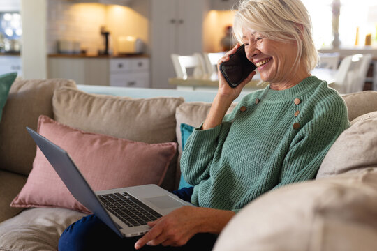 Happy senior caucasian woman sitting on sofa in living room, using laptop and talking on smartphone