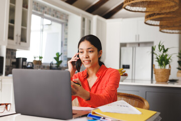 Happy biracial woman sitting at table in kitchen, using laptop and talking on smartphone