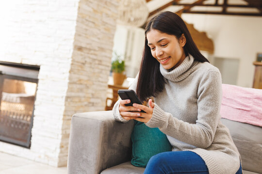 Happy biracial woman sitting on sofa in living room, using smartphone