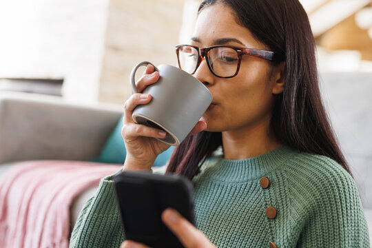 Happy biracial woman sitting on floor in living room, using smartphone and drinking coffee