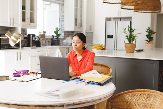Happy biracial woman sitting at table in kitchen, using laptop