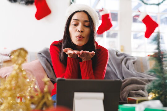 Happy biracial woman wearing santa hat, sitting on sofa in living room, using tablet for video call