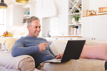 Happy caucasian man sitting on sofa in living room, using laptop for online shopping