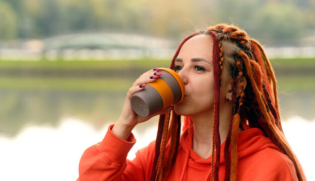 Side view of young woman drinking coffee. Female with redheads dreadlocks with a cup of coffee or tea on background of lake