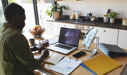 Happy african american man sitting at table in kitchen using laptop