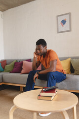 Happy african american man sitting on sofa in living room, talking on smartphone