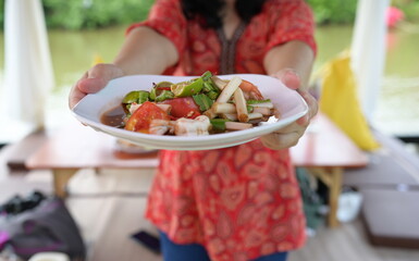 Hand holding plate of food in front to invite you to eat spicy food (Young lotus stems papaya salad with boiled shrimp, pickled fish, lemon, tomato and red peppers).  Woman in red shirt with Thaifood 