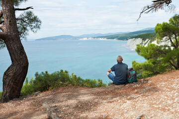 An aged tourist is sitting on a high steep seashore. There is a backpack next to him. In front of him is a delightful landscape: white rocks with pine trees and turquoise water.