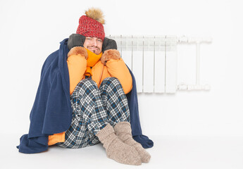 Frightened man feeling cold in hat and down jacket sitting close to radiator. Gas crisis concept