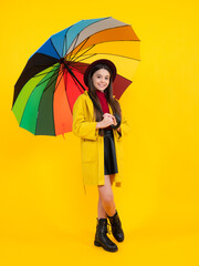 Child girl with rainbow umbrella in autumn weather isolated on yellow background. Autumn kids clothes. Smiling girl in autumn coat. Happy teenager portrait full length.