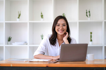 Smiling beautiful young Asian businesswoman sitting with laptop and computer while doing some paperwork at the office. Looking at camera.