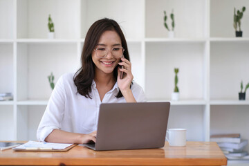 Smiling beautiful young Asian business woman enjoying phone call with customer and commenting at the office while working on a laptop computer.