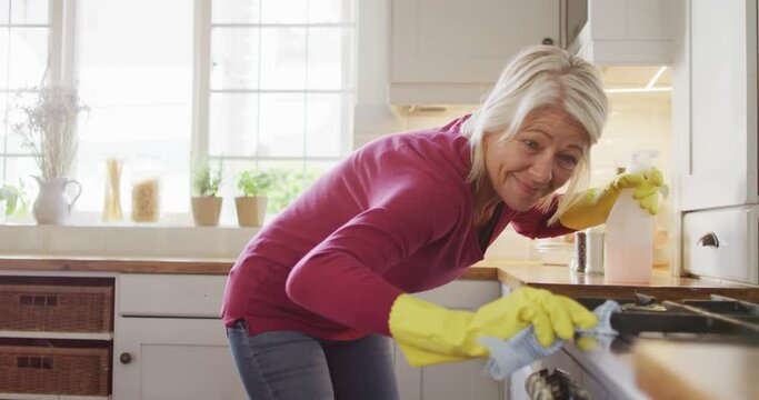 Happy senior caucasian woman wearing rubber gloves, cleaning countertop in kitchen