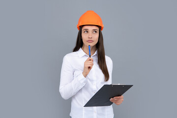 Young woman in hard hat helmet with clipboard isolated on grey background. Architect woman wear helmet and shirt. Thinking architect worker, female constructor engineer.