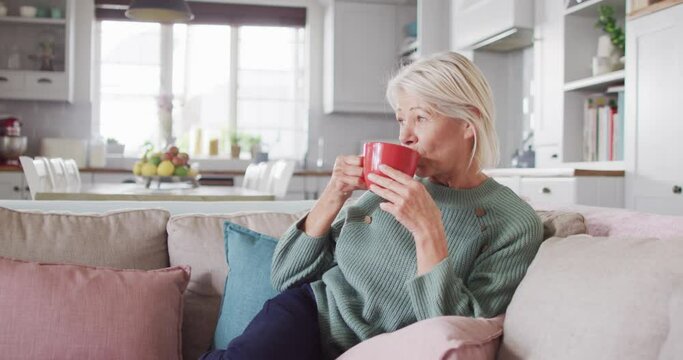 Thoughtful senior caucasian woman sitting on sofa in living room, drinking coffee