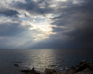 Man standing on cliffs against open sea at evening, storm clouds and sun streaks.