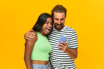 happy multiracial couple surprised looking at one mobile phone, isolated over yellow background.