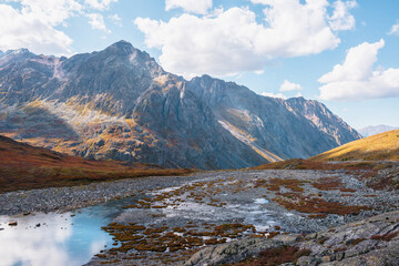Beautiful mountain lake in hanging valley in orange autumn colors with view to large mountain range in sunlight. Lovely autumn landscape with small lake and high mountain top under clouds in blue sky.