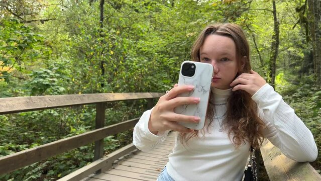 A girl in a white turtleneck takes a selfie holding a phone in front of her in a white case Girl teenager in blue clothes in the park in nature summer Tynehead Regional Park in Surrey