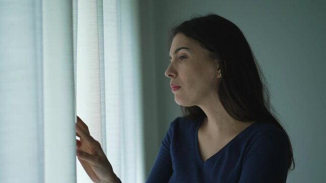 Anxious Woman standing by window peeking out in suspicion feeling distrust