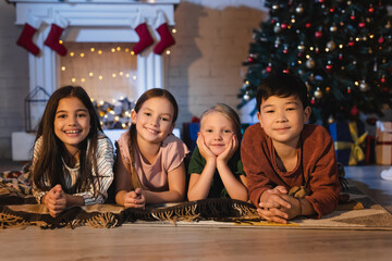 Smiling interracial kids looking at camera while lying on blanket on floor near christmas tree