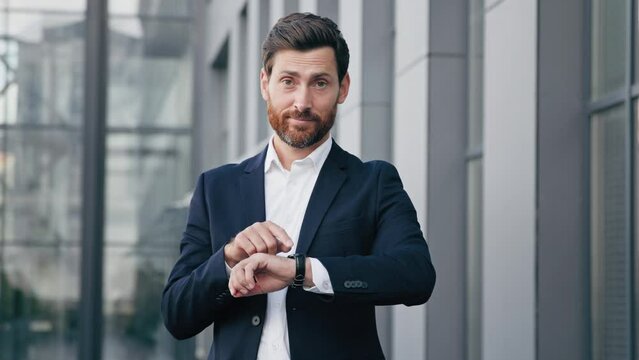 Professional confident businessman standing outdoors waiting for meeting looks at wristwatch checking time displeased caucasian man pointing on watch looking at camera dissatisfied with being late