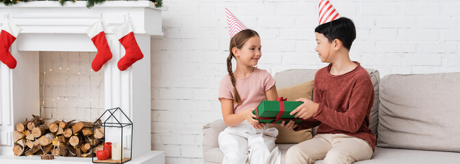 Asian boy in party cap giving present to friend during birthday party near christmas decor at home, banner