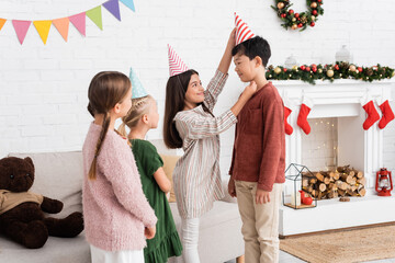 Obraz premium Smiling girl wearing party cap on asian friend during birthday party and christmas celebration