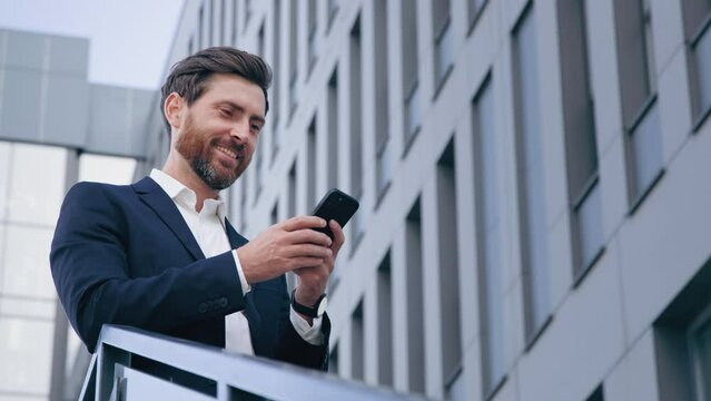 Stylish handsome businessman holding phone reading email stands on street smiling writes message communicates in social network pensive man browses goods in online store using mobile app on smartphone