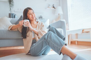 Happy young asian woman drinking coffee relaxing on the rug beside to the sofa at home. Smiling female enjoying resting sitting on couch in modern living room. - Powered by Adobe