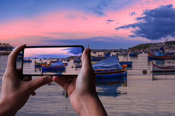 Tourist taking photo of traditional Maltese boats in Marsaxlokk harbor during sunset, Malta