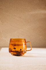 Glass transparent orange mug with hot green tea, shadow and reflection on light stone background. Front view