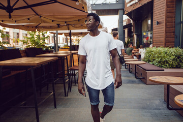 An african american man in a white t-shirt stands in a street cafe. Mock-up.