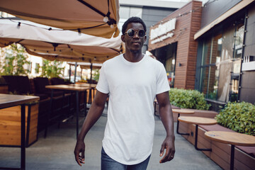 Fototapeta premium An african american man in a white t-shirt stands in a street cafe. Mock-up.
