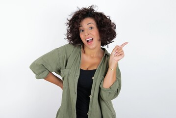 young beautiful woman with curly short hair wearing green overshirt over white wall holding finger up having idea and posing