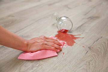 Glass of red wine fell on laminate, wine spilled on floor. A woman wipes the laminate from moisture.