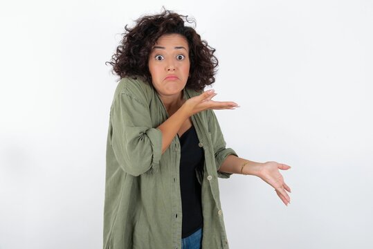 young beautiful woman with curly short hair wearing green overshirt over white wa pointing aside with both hands showing something strange and saying: I don't know what is this. Advertisement concept.