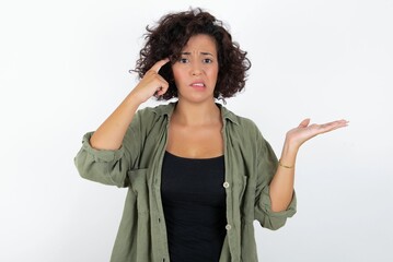 young beautiful woman with curly short hair wearing green overshirt over white wall confused and annoyed with open palm showing copy space and pointing finger to forehead. Think about it.