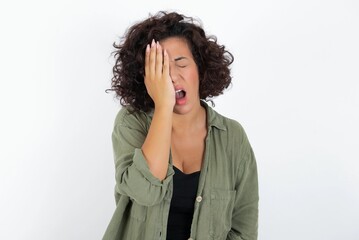 young beautiful woman with curly short hair wearing green overshirt over white wall Yawning tired...