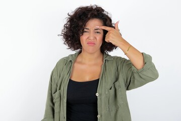 young beautiful woman with curly short hair wearing green overshirt over white wall pointing...