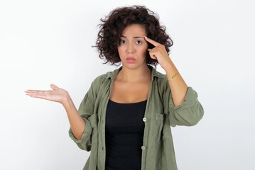 young beautiful woman with curly short hair wearing green overshirt over white wall confused and...