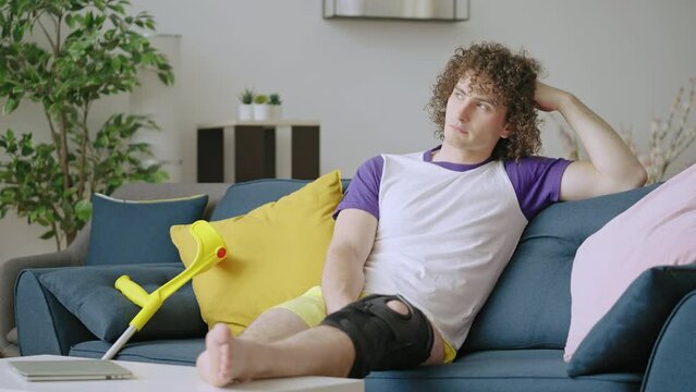 Unhappy injured young man sitting on couch, bandage on leg, knee sprain trauma