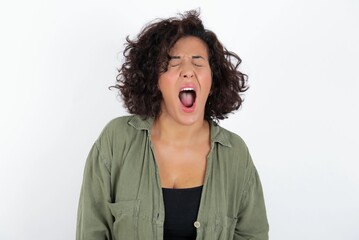 young beautiful woman with curly short hair wearing green overshirt over white wall angry and mad screaming frustrated and furious, shouting with anger. Rage and aggressive concept.