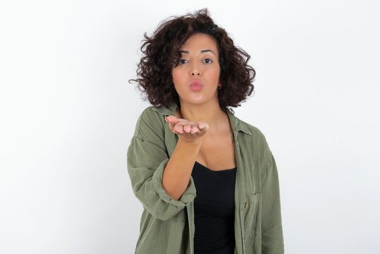 young beautiful woman with curly short hair wearing green overshirt over white wall looking at the camera blowing a kiss with hand on air being lovely and sexy. Love expression.