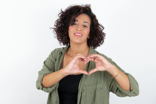 young beautiful woman with curly short hair wearing green overshirt over white wall smiling in love doing heart symbol shape with hands. Romantic concept.