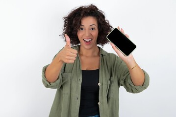 Portrait young beautiful woman with curly short hair wearing green overshirt over white wall holding in hands cell showing giving black screen thumb up