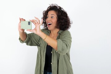young beautiful woman with curly short hair wearing green overshirt over white wall taking a selfie...