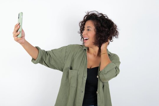 young beautiful woman with curly short hair wearing green overshirt over white wall smiling and taking a selfie ready to post it on her social media.