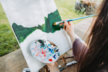 Unrecognizable young female artist painting her own mountain scenery with oil on a canvas. Amateur...